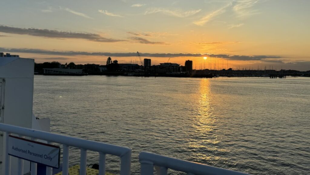 The photo captures a tranquil sunset over Gosport's waterfront. The sky is painted with soft hues of orange, yellow, and blue as the sun descends towards the horizon, casting a golden reflection on the water's surface. In the distance, silhouettes of buildings, cranes, and masts of boats can be seen against the bright sky. 