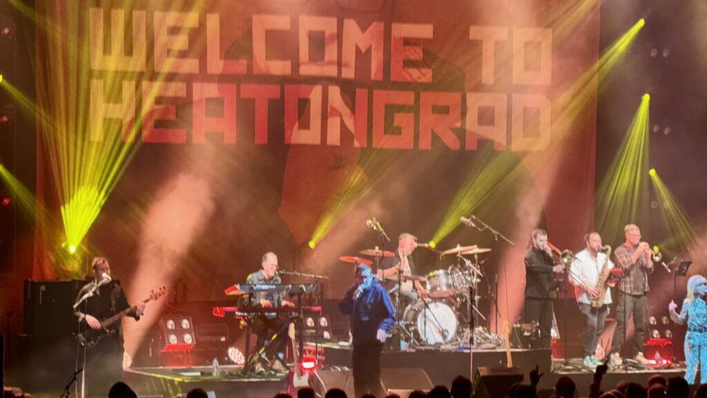 This image depicts a lively musical performance on a stage with dramatic lighting effects. The background features a large banner with bold red text that says, "WELCOME TO HEATONGRAD." Yellow and green beams of light pierce through haze or stage smoke, creating a dynamic atmosphere. On stage, multiple musicians are performing.