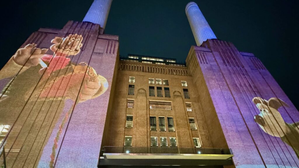 This is an image of Battersea Power Station in London at night, featuring a vibrant projection of Wallace and Gromit, iconic characters from the beloved British animated series. The projection is cast onto the station's façade, with the tall chimneys visible above, illuminated in soft light.