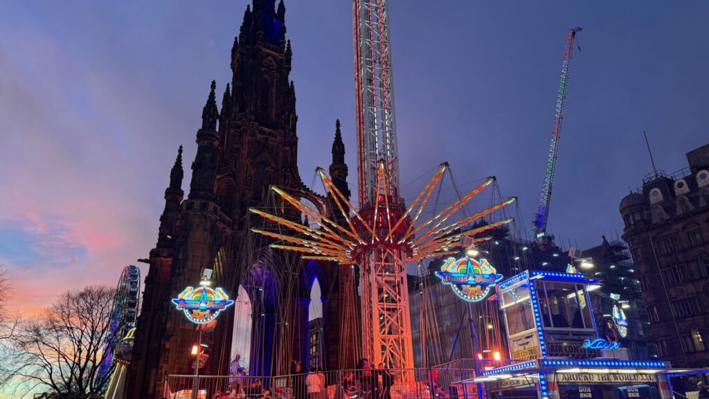 This image captures the festive atmosphere in Edinburgh on Christmas Eve. The scene features the Scott Monument illuminated against the evening sky, with its Gothic architecture beautifully lit. In the foreground, a vibrant fairground attraction, Star Flyer, stands out with colourful lights and intricate design, adding energy to the scene. A Ferris wheel is visible on the left, glowing against the soft hues of a wintry sunset.