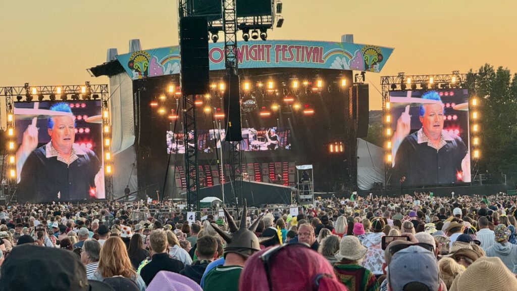 This image shows a large outdoor festival stage with two large video screens displaying the band, Green Day The stage has a colourful "ISLE OF WIGHT FESTIVAL" banner at the top and is lit with numerous bright stage lights. The crowd is very dense, with festivalgoers wearing various hats and summer clothing. The photo appears to be taken at sunset or dusk, giving the sky a golden hue.