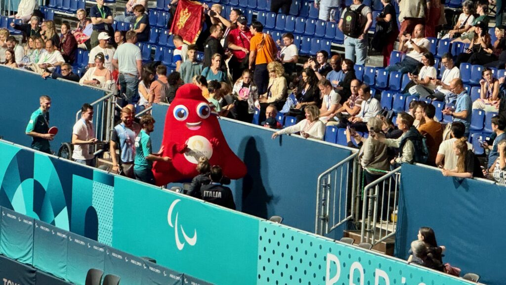 This image shows a sports venue with Paralympic Games branding visible on turquoise barriers with the Paralympic symbol. The main focus is a large red mascot character with big blue eyes and a friendly smile, likely Phryge, the official mascot of Paris 2024. The mascot is interacting with spectators in stadium seating filled with a diverse crowd.