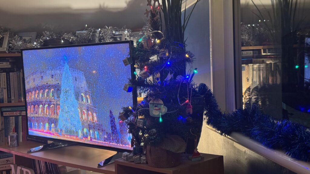 The image shows a small Christmas tree decorated with coloured lights and ornaments, positioned next to a TV displaying what the Colosseum in Rome lit up at night. Silver tinsel garland is visible along the top edge of the frame, and there bookshelves in the background.