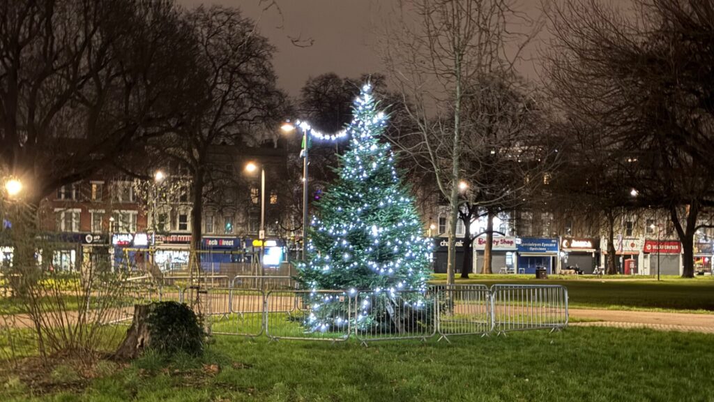 A night-time photo of a Christmas tree on Shepherd's Bush Green in London, taken in mid-January. The tree is decorated with bright white LED lights and is surrounded by temporary metal barriers. The scene captures that particular post-Christmas period in London when some festive decorations remain up into the new year, creating a lingering sense of the holiday season even as winter properly sets in.