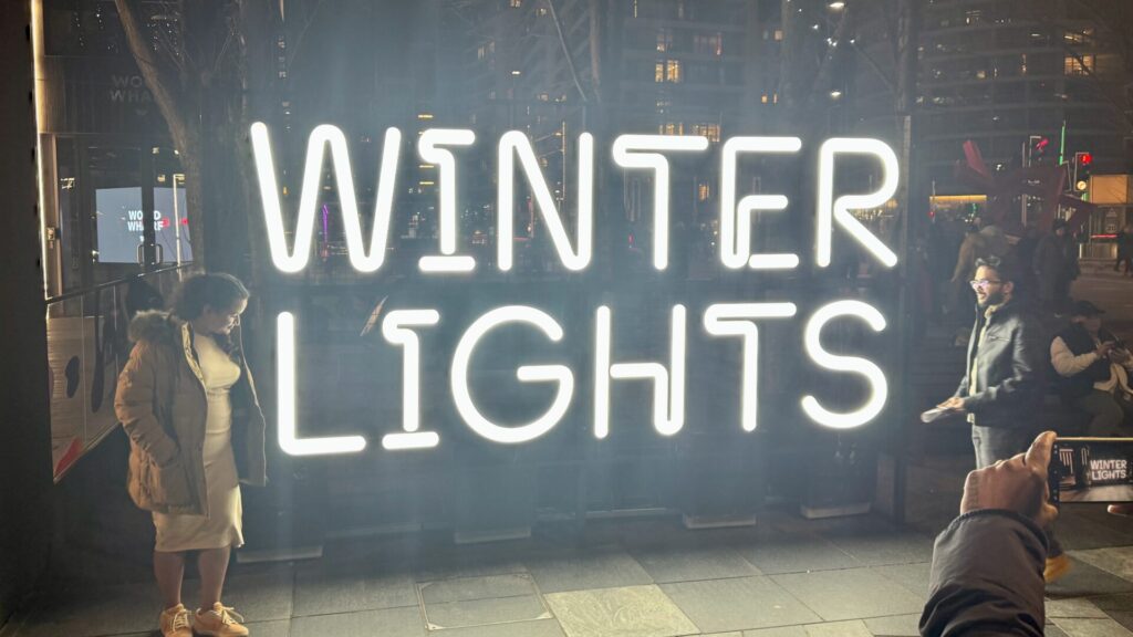 This image shows a large, illuminated "WINTER LIGHTS" sign in bright white neon lighting, creating a striking display against the dark evening backdrop. The sign is part of the Canary Wharf Winter Light festival installation. Several people can be seen around the sign, including some taking photos of the installation. The scene has a misty, atmospheric quality, with the lights from surrounding buildings and the illuminated sign creating a warm glow in the winter evening.