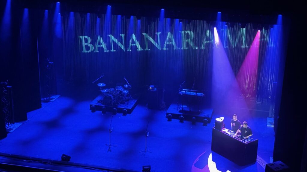 The image shows a theatre stage illuminated in deep blue lighting, with "BANANARAMA" visible in turquoise lettering above. The stage is set up with a drum kit on a riser in the centre background and various other instruments and equipment positioned around the stage. Two DJs are visible at a control desk on the right side, lit by purple and pink spotlights. The theatre's curtains create a dramatic backdrop with vertical striping effects from the lighting.