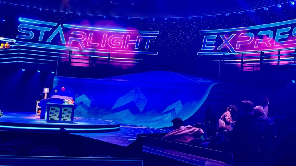 The image shows a theatrical stage for "Starlight Express" illuminated in blue and pink neon lighting. The title "STARLIGHT EXPRESS" appears in bright neon lettering across the top. The set features curved racing tracks and geometric mountain shapes in the background. The lighting creates a starry night effect, and some audience members are visible in the foreground.