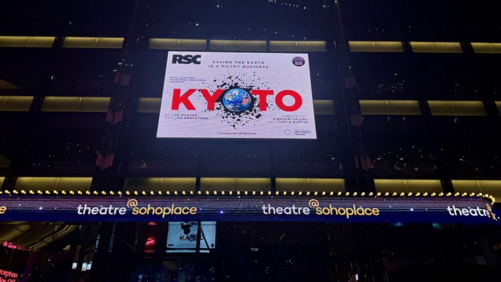 This image shows the exterior display of Sohoplace theatre at night, featuring a digital billboard for a play titled "KYOTO". The billboard includes the RSC (Royal Shakespeare Company) logo and the tagline "SAVING THE EARTH IS A FILTHY BUSINESS". The show's title is displayed in large red letters with an image of Earth in the middle of the 'O', surrounded by what appears to be splatter or debris effects.